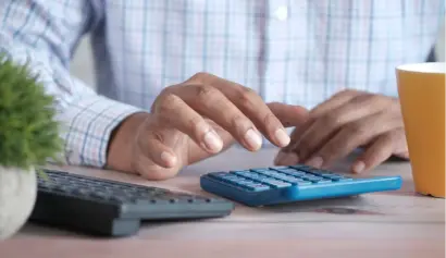 A person using a calculator next to a keyboard and a coffee cup on a wooden desk