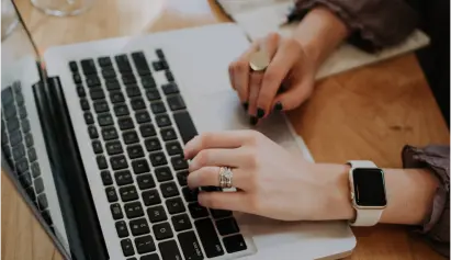 Person typing on laptop with smartwatch and rings, notebook in background
